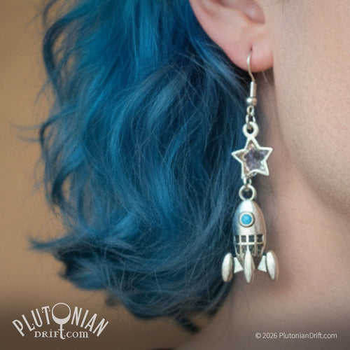 As Astra - retro rocketship earrings. A model with blue hair wears a silver rocketship earring. A retro ship with a turquoise porthole dangles below a crystal star. Plutonian Drift.