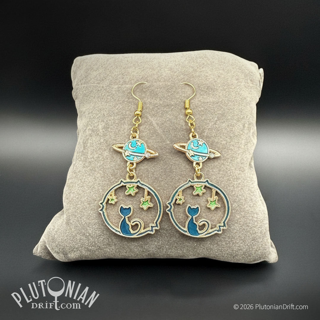The Catsronaut space cat celestial earrings on a grey velvet pillow and grey background. A blue and green enamel cat framed by a hoop and stars hangs below a blue and green ringed planet. Gold tone fittings. Plutonian Drift.