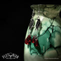 Pair of black enamel crow earrings with silver hooks and chains, deep red teardrop drops, displayed on a green cracked ceramic form by Plutonian Drift.