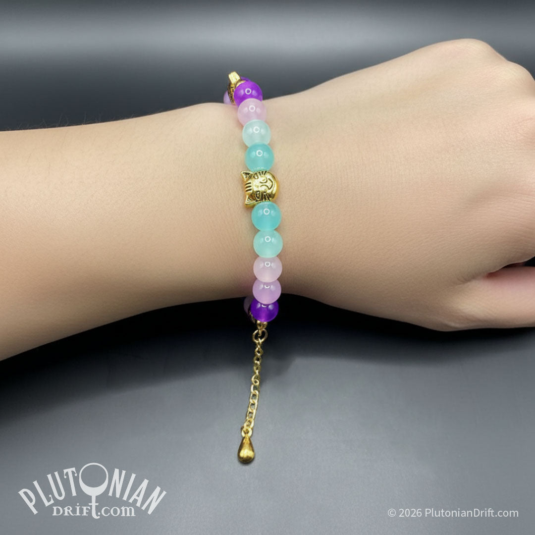 Nothing But the Grin - Alice in Wonderland inspired beaded bracelet. Violet, pink and turquoise glass beads with antique gold-tone grinning cat head beads on a model's wrist. Plutonian Drift.