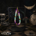 Nothing But the Grin - Alice in Wonderland inspired beaded bracelet. Violet, pink and turquoise glass beads with antique gold-tone grinning cat head beads hanging on a stand on a chequered table. Clocks, teacups and mushrooms surround it and a disembodied grin floats behind. Plutonian Drift.