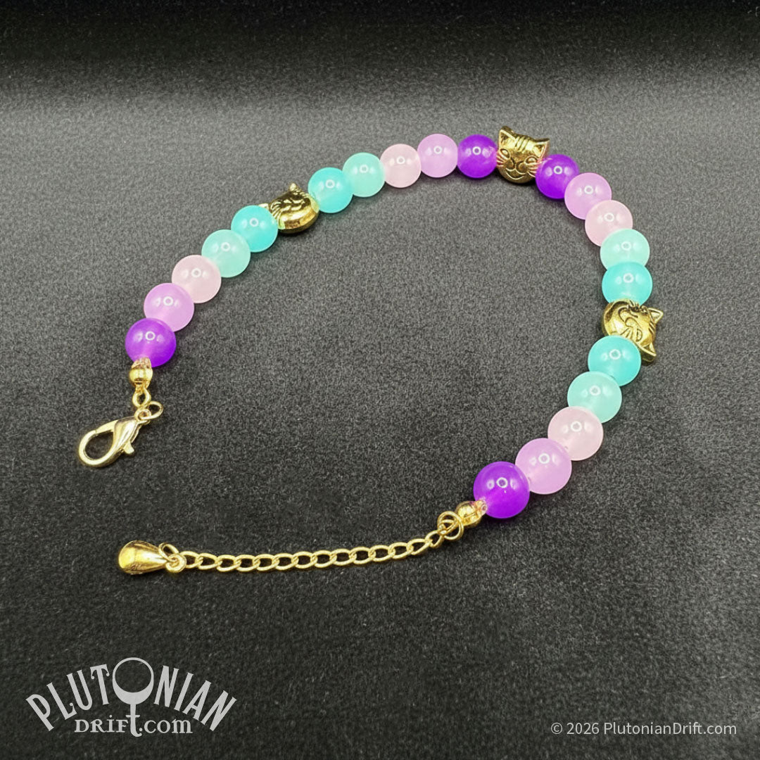 Nothing But the Grin - Alice in Wonderland inspired beaded bracelet. Violet, pink and turquoise glass beads with antique gold-tone grinning cat head beads on a grey velvet background. Plutonian Drift.