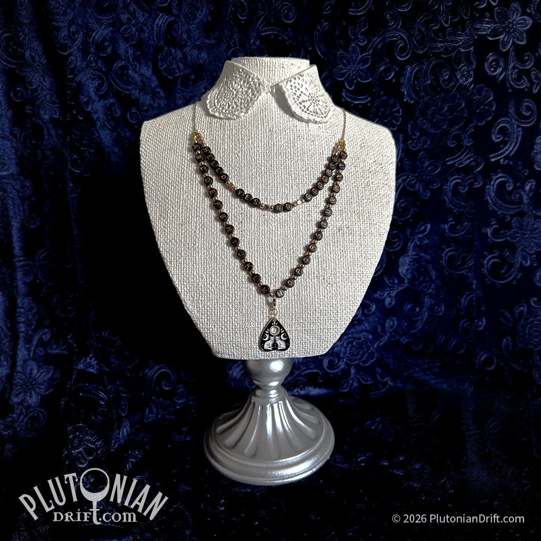 Hello/Goodbye ouija necklace. A draping necklace layed out like a ouija board rests on a white necklace stand against a blue velvet backdrop. Black and gold letter beads spell out hello, yes, no, goodbye on one row, and the alphabet on the second row. A gold planchette charm with two white enamel cats is clipped in the center of the lower row. Plutonian Drift.