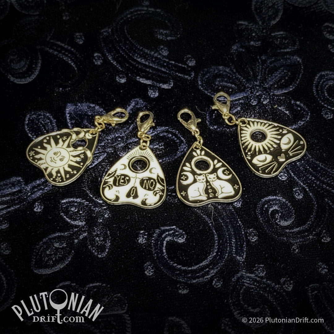 Hello/Goodby ouija necklace. Four detachable gold charms with black and white enamel on a blue velvet cloth. From left to right: sun design, skull with Yes in one eye and No in the other, two sitting white cats with moons, a black cat face below a sun. Plutonian Drift.