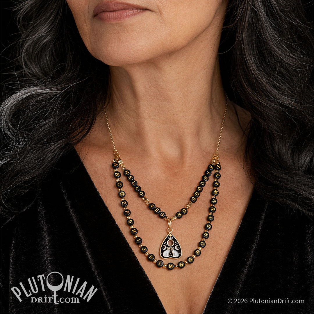 Hello/Goodbye ouija necklace. An older woman with dark hair wears a draping necklace layed out like a ouija board. Black and gold letter beads spell out hello, yes, no, goodbye on one row, and the alphabet on the second row. A gold planchette charm with two white enamel cats is clipped in the center of the top row. Plutonian Drift.