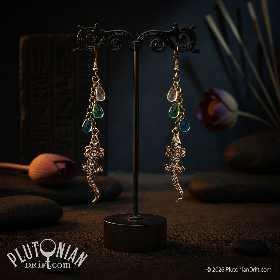 Pair of gold crocodile earrings with green glass teardrops on a stand on a dark sandbank.