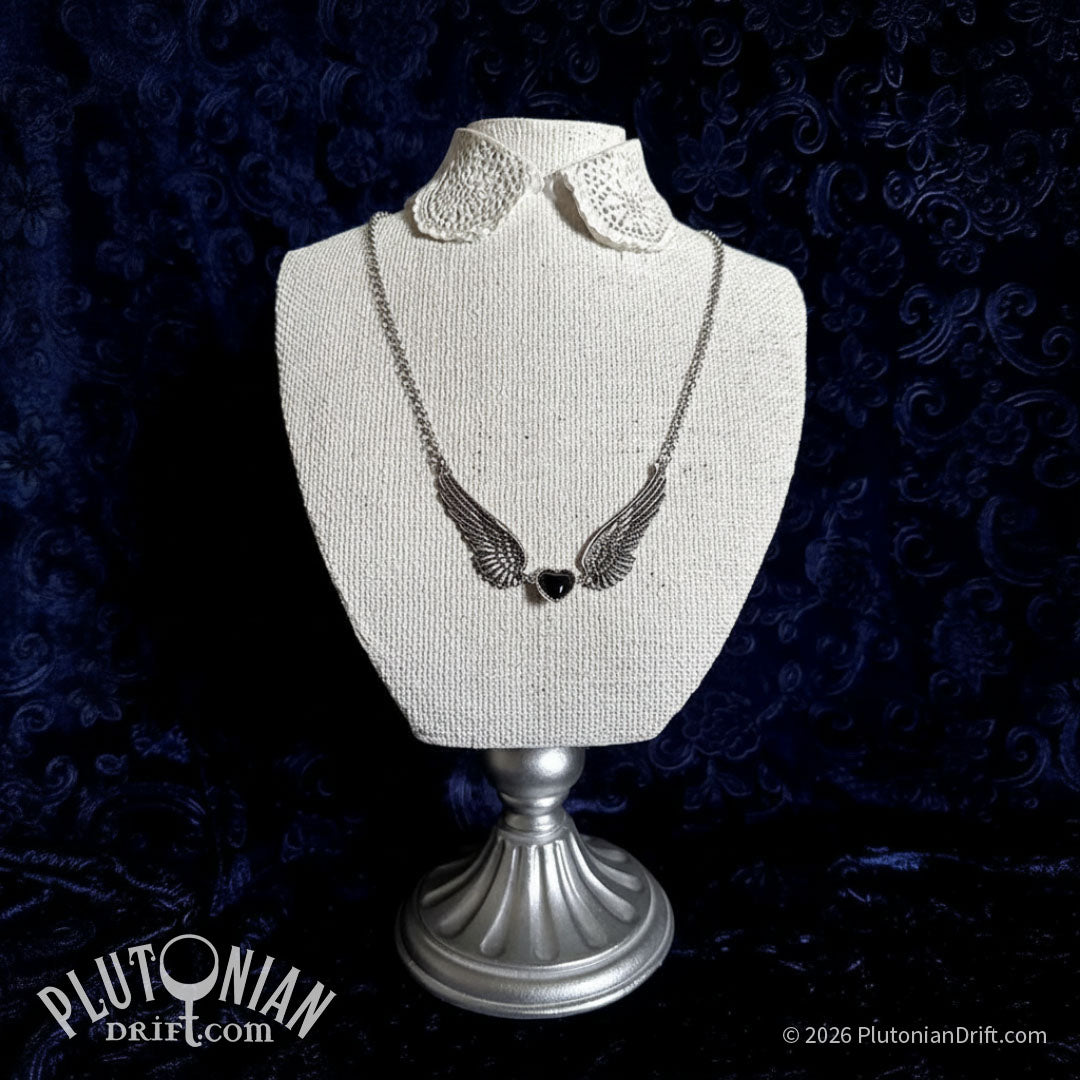 Sepulchre-by-the-Sea statement necklace on a white stand against a blue velve backdrop. A black glass heart in silver-toned setting sits between two outstretched zinc alloy angel wing on a silver-toned chain. 4.75 inch wingspan. Plutonian Drift.