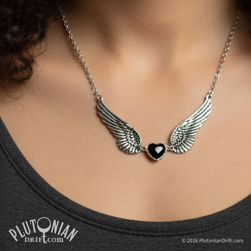 Sepulchre-by-the-Sea statement necklace worn by a model. A black glass heart in silver-toned setting sits between two outstretched zinc alloy angel wing on a silver-toned chain. 4.75 inch wingspan. Plutonian Drift.