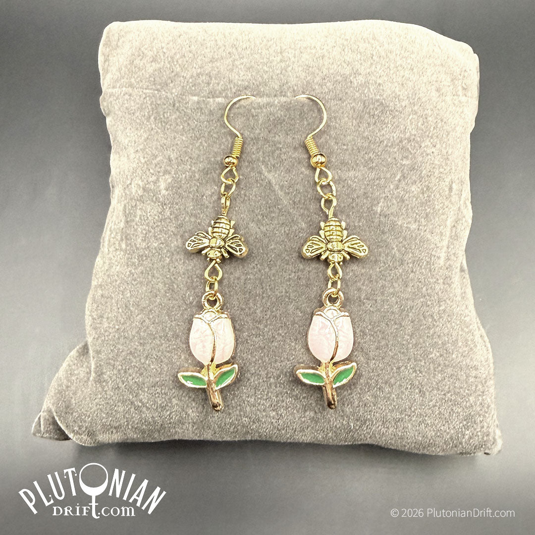 Sweet Industry - botanical honeybee and tulip earrings. Earrings that feature an antique gold tone bee dangling above a light pink enamel tulip charm rest on a grey velvet pillow. Plutonian Drift.