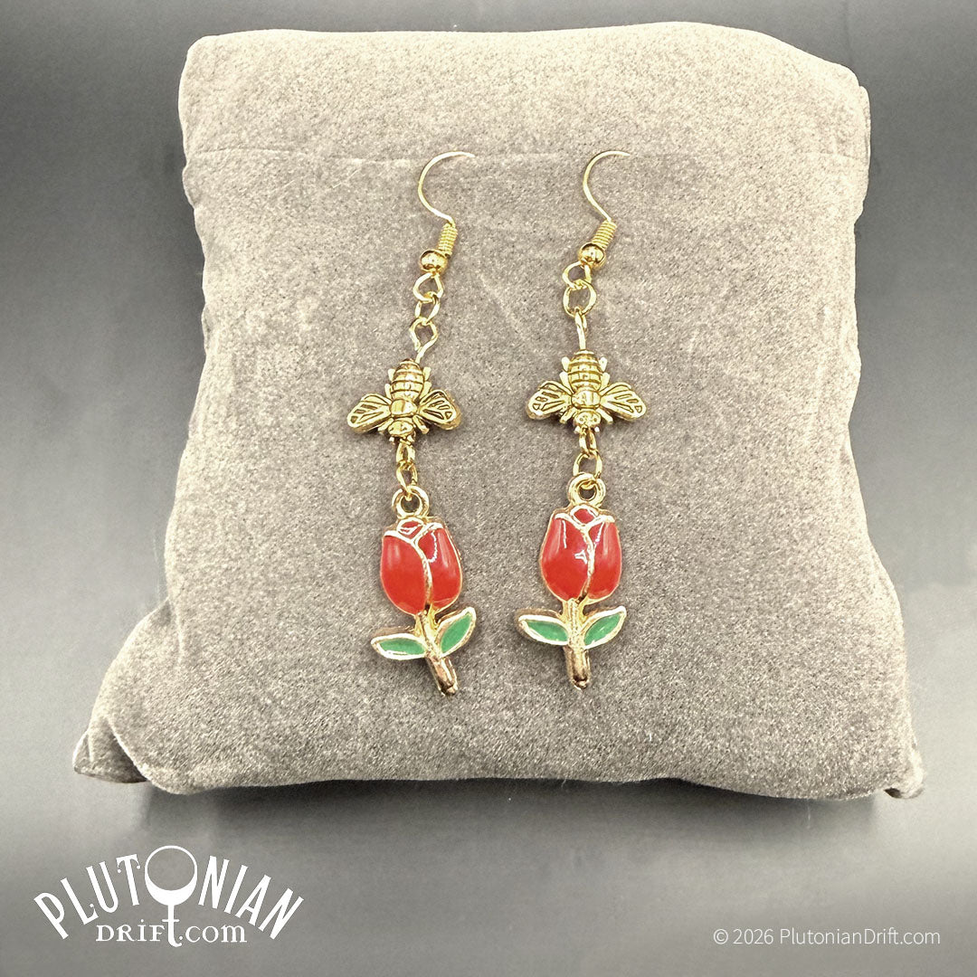 Sweet Industry - botanical honeybee and tulip earrings. Earrings that feature an antique gold tone bee dangling above a red enamel tulip charm rest on a grey velvet pillow. Plutonian Drift.