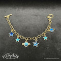 Travel Guide - Galactic tour charm bracelet. A gold tone chain charm bracelet against a grey velvet background. Charms are blue enamel stars and ringed planets. Plutonian Drift.