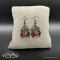 Pair of black gothic spider earrings with faceted red bodies on a beige display cushion, from Plutonian Drift in Fables and Shop Earrings