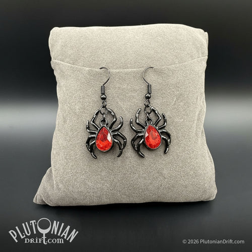 Pair of black gothic spider earrings with faceted red bodies on a beige display cushion, from Plutonian Drift in Fables and Shop Earrings