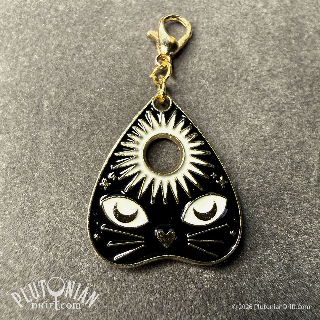 Hello/Goodby ouija necklace. A detachable gold planchette charm with black and white enamel on a grey velvet cloth. Option 3: a black cat face below a sun. Plutonian Drift.