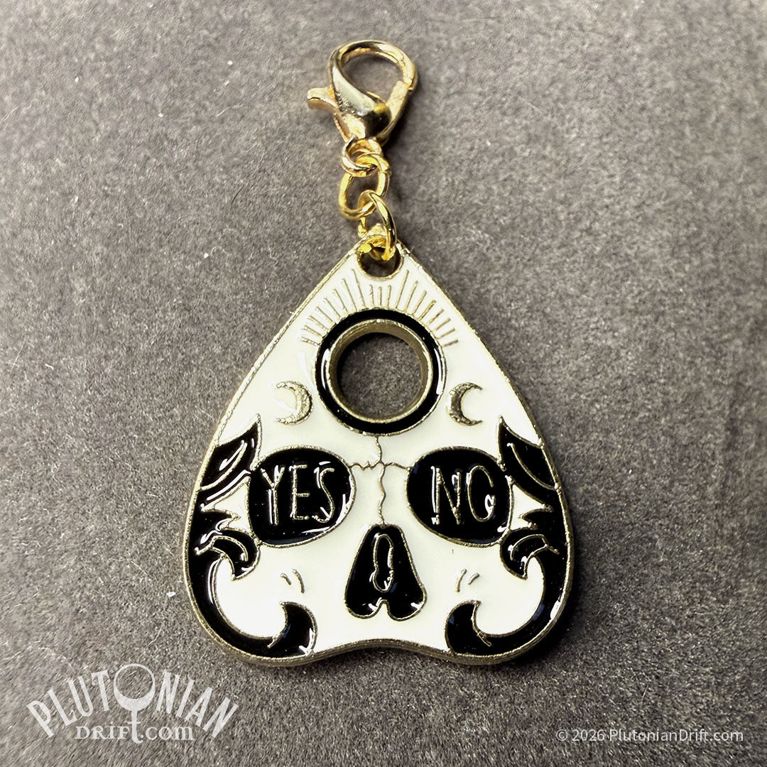 Hello/Goodby ouija necklace. A detachable gold planchette charm with black and white enamel on a grey velvet cloth. Option 2: skull design with the word Yes in one eye and No in the other. Plutonian Drift.