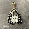 Hello/Goodby ouija necklace. A detachable gold planchette charm with black and white enamel on a grey velvet cloth. Option 1: Sun design with face and rays. Plutonian Drift.