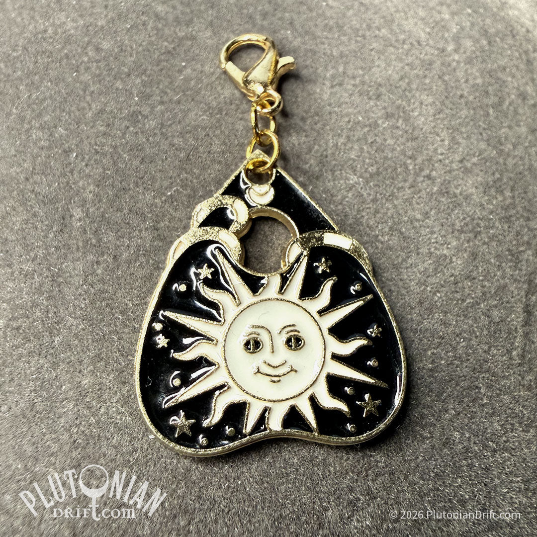 Hello/Goodby ouija necklace. A detachable gold planchette charm with black and white enamel on a grey velvet cloth. Option 1: Sun design with face and rays. Plutonian Drift.